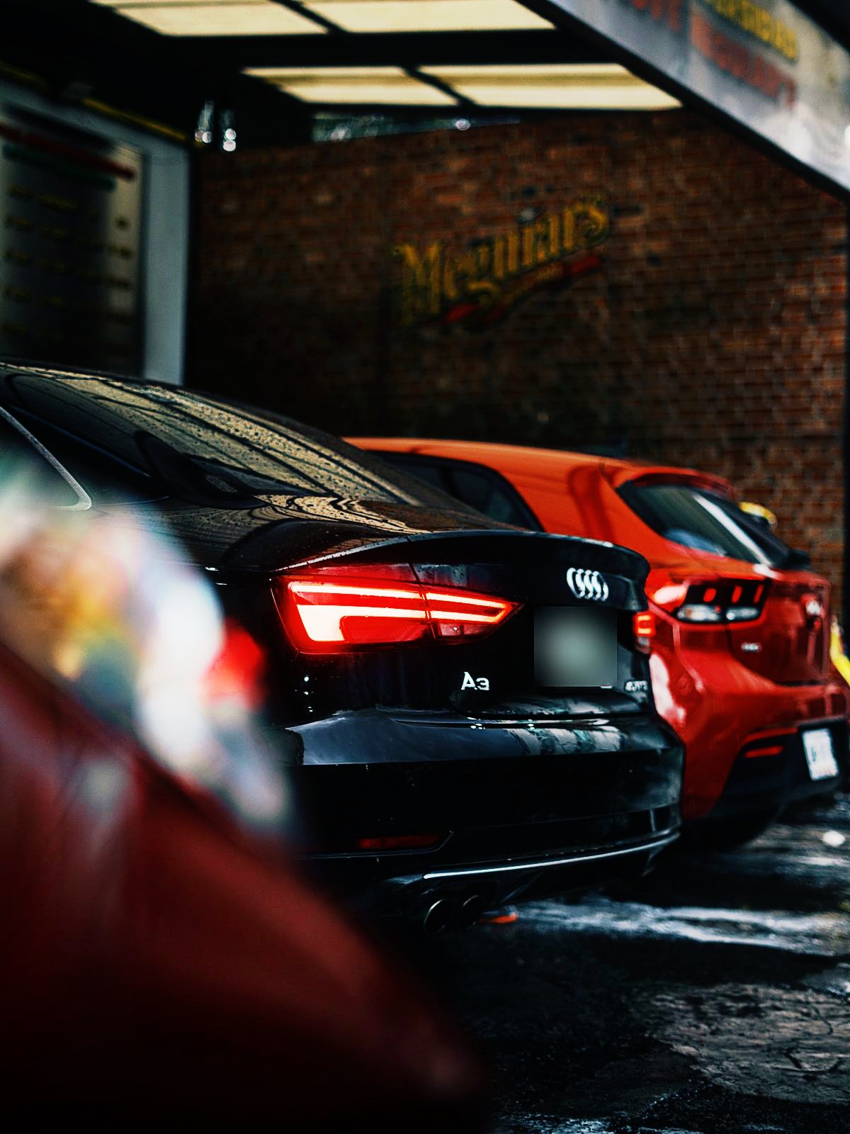 Audi A3 negro y un auto rojo recién lavados frente al logo de Meguiar’s en la pared de ladrillo